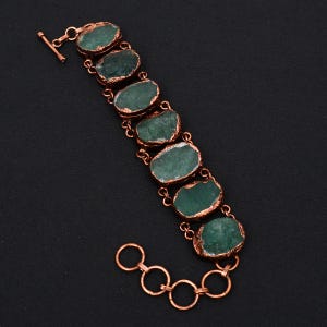 Puede incluir: Un brazalete de cobre con cierre de palanca presenta siete cuentas de piedras preciosas verdes. Las cuentas tienen una forma irregular y una textura rugosa y natural.