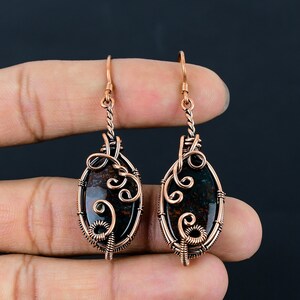 May include: A pair of copper wire wrapped earrings with oval shaped green and brown jasper stones. The earrings have a delicate, intricate design.