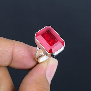 May include: A silver ring featuring a large, rectangular, faceted red gemstone set in a polished metal bezel. The stone has a deep crimson hue with visible internal clarity and light reflection across its flat, angled top surface.