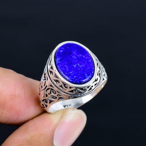 May include: A silver ring with an oval, deep blue gemstone. The ring has an ornate, patterned design around the gemstone and on the band. The ring is held up by a person's fingers against a dark background.