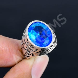 May include: A silver-toned ring featuring a large, oval-shaped, faceted blue gemstone set in a band with intricate, openwork scroll patterns. The metal band displays a dark, oxidized finish within the recessed areas of the decorative metalwork.