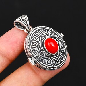 May include: A round silver locket pendant featuring an intricate embossed scrollwork pattern and a central oval-shaped smooth red stone. The pendant includes a decorative triangular bail at the top and a visible hinge mechanism on the side.