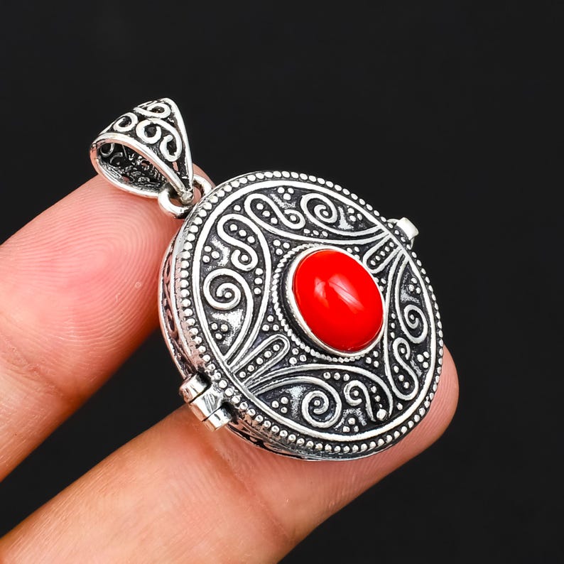 May include: A round silver locket pendant featuring an intricate embossed scrollwork pattern and a central oval-shaped smooth red stone. The pendant includes a decorative triangular bail at the top and a visible hinge mechanism on the side.
