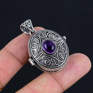 May include: An oval-shaped silver locket pendant featuring an intricate embossed scrollwork pattern and a central polished purple amethyst gemstone. The pendant includes a decorative silver bail at the top and a hinged side mechanism for opening.