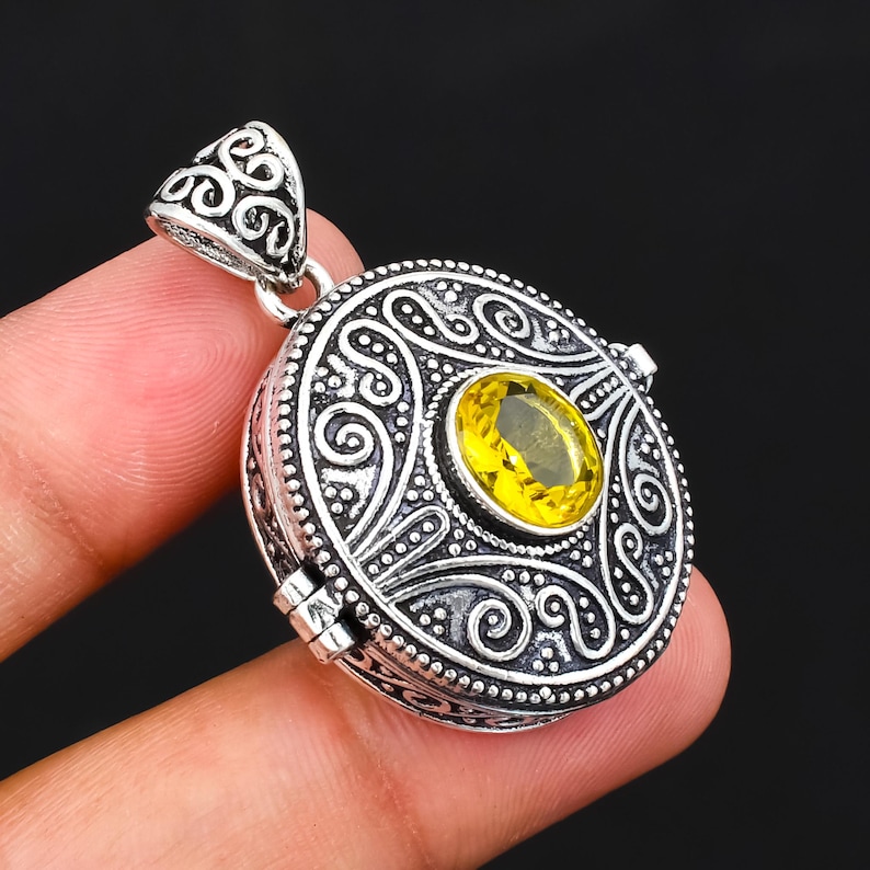 May include: A round silver locket pendant featuring an oval-cut yellow gemstone at the centre. The metal surface displays intricate embossed scrollwork and oxidised detailing, with a decorative filigree bail at the top for chain attachment.