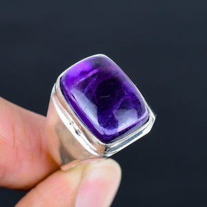 May include: A silver ring featuring a large, rectangular, cabochon-cut purple amethyst gemstone. The stone is set in a smooth, polished silver bezel that transitions into a wide, solid silver band with a clean, reflective metallic finish.