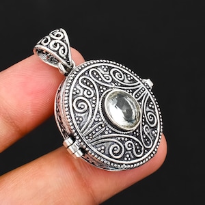 May include: A round silver pendant featuring an intricate embossed scrollwork pattern and a central clear faceted gemstone. The pendant includes a decorative triangular bail at the top and a visible hinge mechanism on the side of the circular frame.