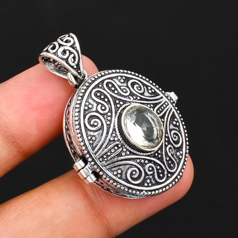 May include: A round silver pendant featuring an intricate embossed scrollwork pattern and a central clear faceted gemstone. The pendant includes a decorative triangular bail at the top and a visible hinge mechanism on the side of the circular frame.