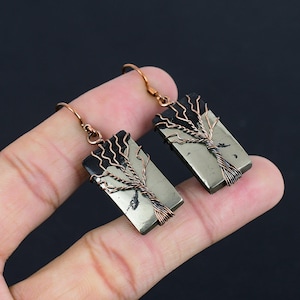 Può includere: Un paio di orecchini con una pietra di pirite rettangolare con un design ad albero di filo di rame. La pietra è di colore marrone chiaro con venature nere.