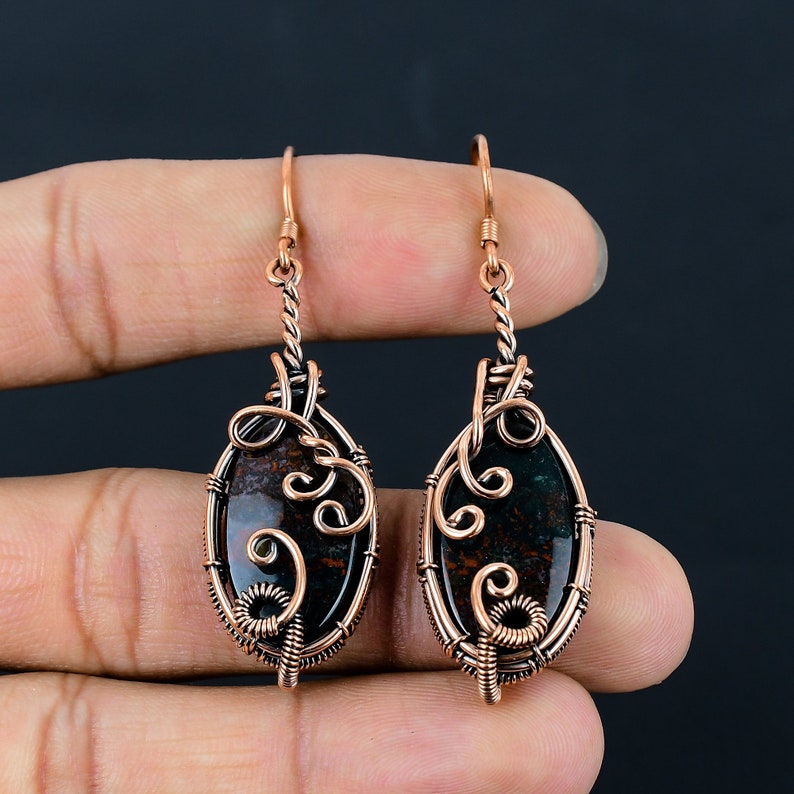 May include: A pair of copper wire wrapped earrings with oval shaped green and brown jasper stones. The earrings have a delicate, intricate design.