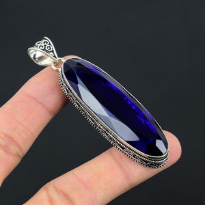 Puede incluir: Un colgante largo de forma ovalada con una piedra preciosa facetada de color azul intenso, engastada en un marco decorativo de plata de ley con intrincados detalles metálicos en los bordes y una anilla de plata a juego en la parte superior.
