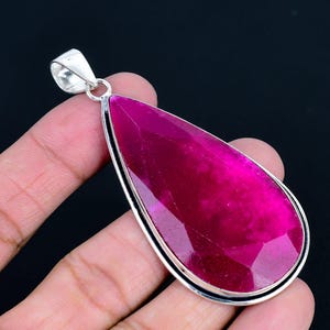 May include: A teardrop-shaped pendant featuring a large, faceted, vibrant magenta gemstone set in a polished silver-toned metal frame with a matching silver-toned bail at the top for attachment to a chain or cord.