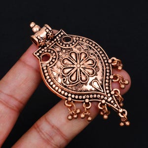 Puede incluir: Un colgante de metal en tono cobre con una forma de lágrima simétrica y ornamentada, que presenta un motivo floral central, patrones en relieve intrincados, pequeños dijes esféricos colgantes en los bordes inferiores y una reasa cilíndrica.