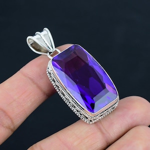 May include: A rectangular pendant featuring a large, faceted deep purple gemstone set in a silver frame with intricate filigree detailing along the sides. The pendant includes a triangular silver bail at the top for attachment to a chain or cord.