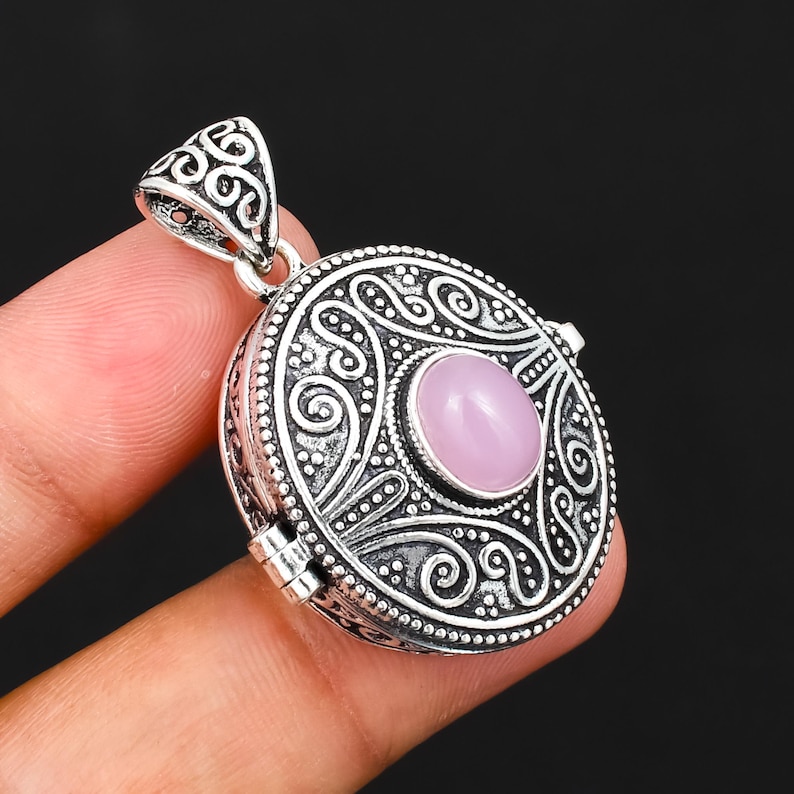 May include: A round silver locket pendant featuring an intricate embossed scrollwork pattern and a central oval cabochon of smooth, pale pink rose quartz. The metal has an oxidised finish to highlight the detailed texture and decorative metalwork.