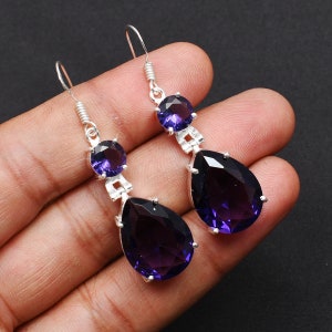 May include: A pair of silver dangle earrings with teardrop-shaped amethyst gemstones. The earrings have a smaller round amethyst gemstone above the teardrop.