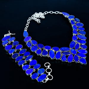 Puede incluir: Un conjunto de joyas que incluye un collar, una pulsera y pendientes. El conjunto está hecho de piedras preciosas pulidas de color azul oscuro engastadas en metal plateado. El collar y la pulsera tienen un diseño en capas.