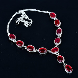 Op de afbeelding: Een zilveren ketting met een reeks gefacetteerde, ovaalvormige dieprode stenen in individuele metalen zettingen. De ketting heeft fijne schakels met een haaksluiting en een centraal hangend gedeelte met twee extra stenen.