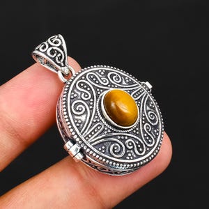 May include: A round silver pendant featuring an oval tiger's eye gemstone at the centre. The metal surface is decorated with intricate embossed scrollwork and swirling patterns. A matching silver bail with a cutout design is attached to the top.