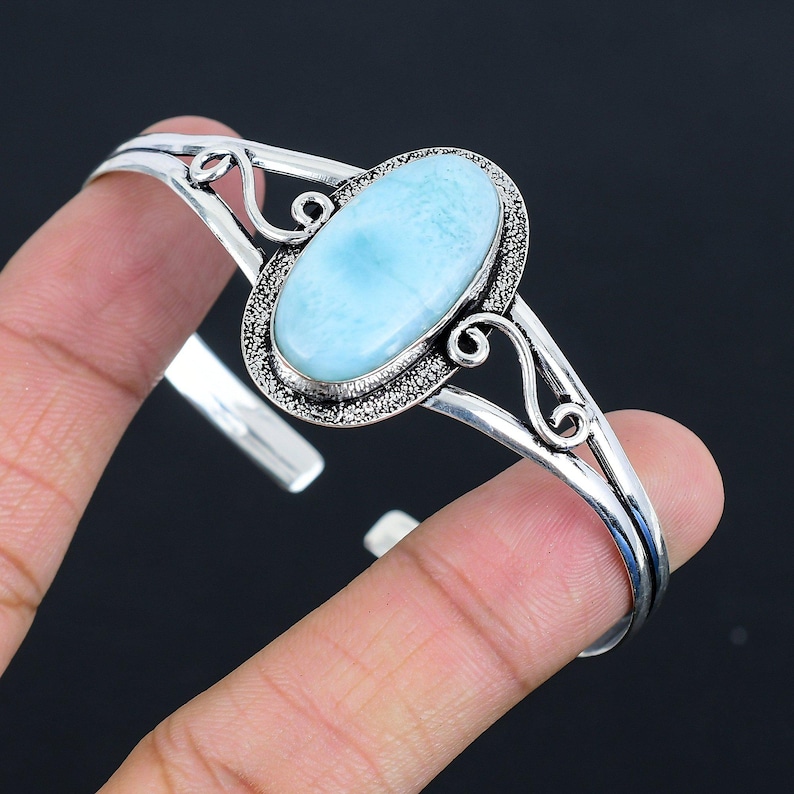 May include: A silver bangle bracelet with a light blue oval cabochon gemstone. The bracelet has a delicate, ornate design.