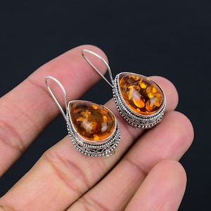 Può includere: Un paio di orecchini pendenti in argento con pietre di ambra a forma di goccia. Le pietre sono di un colore arancione intenso e hanno un aspetto leggermente nuvoloso. Gli orecchini hanno un design delicato e ornato.