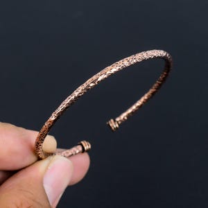 May include: A copper-toned metal cuff bracelet featuring a textured, hammered surface throughout the band. The bracelet has an open design with two small, ribbed cylindrical caps at each end, providing a distinct finish to the metal structure.