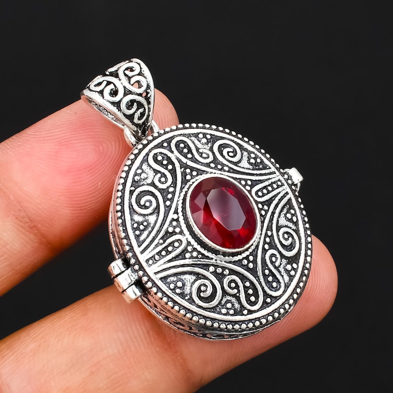 May include: A round silver locket pendant featuring an oval-cut deep red gemstone at the centre. The metal surface is decorated with intricate embossed scrollwork and swirling patterns, finished with a matching silver bail at the top.