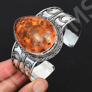 Peut inclure: Un large bracelet manchette orné d'une grande pierre d'ambre orange translucide en forme de poire, sertie sur une bande en métal argenté. La bande métallique présente des motifs de volutes en relief et une bordure perlée autour de la pierre.