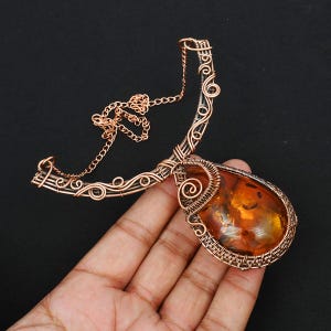 Op de afbeelding: Een ketting met een amberen hanger die is gewikkeld in koperdraad en een delicate ketting heeft. De hanger is druppelvormig en de draad is gewikkeld in een gedetailleerd patroon.