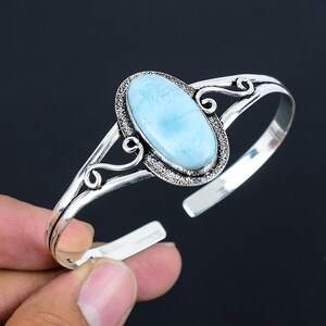 May include: A silver bangle bracelet with an oval-shaped blue gemstone set in a silver bezel. The bracelet has a delicate, ornate design.