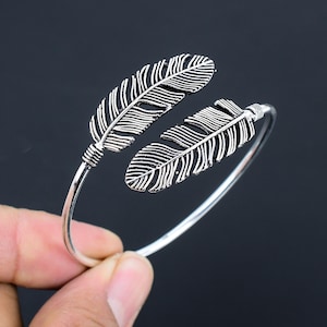 May include: A silver bangle bracelet with two feather charms. The feathers are detailed with lines and are attached to the bracelet with a wire.