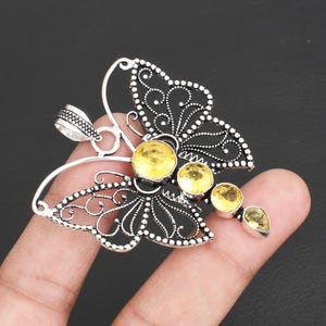 May include: A silver butterfly pendant with detailed filigree wings and yellow gemstone accents. The pendant has a central cluster of round gemstones and a teardrop-shaped stone at the tip. The pendant is held by a finger.