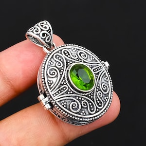 May include: A round silver pendant featuring an intricate embossed scrollwork pattern and a central oval-shaped faceted green peridot gemstone. The pendant includes a decorative bail at the top and a visible hinge mechanism on the side of the frame.