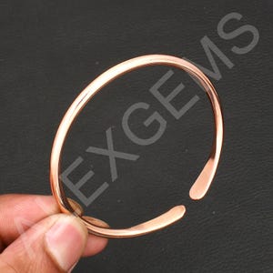 Może przedstawiać: Otwarta bransoletka w kolorze miedzi. Bransoletka to prosta, wypolerowana opaska z zaokrąglonymi krawędziami i niewielką szczeliną na otwarciu. Metal ma ciepły, czerwonobrązowy odcień.