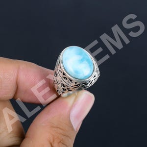 May include: A silver ring featuring a large, oval-shaped, light blue Larimar gemstone with white marbling. The metal band displays an intricate, openwork filigree pattern. The text ALE GEMS is visible in the background of the image.