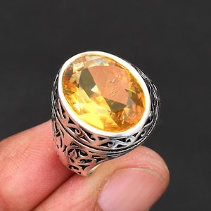 Op de afbeelding: Een zilveren ring met een ovale, gele edelsteen. De ring heeft een sierlijke zilveren band met gedetailleerde patronen. Deze statement ring is geschikt voor speciale gelegenheden.