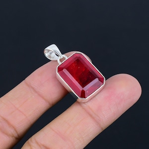 May include: A rectangular pendant featuring a deep red, faceted gemstone set in a polished silver frame. The stone has a clear, glassy texture with sharp, beveled edges. A silver bail is attached to the top of the setting for hanging on a chain.