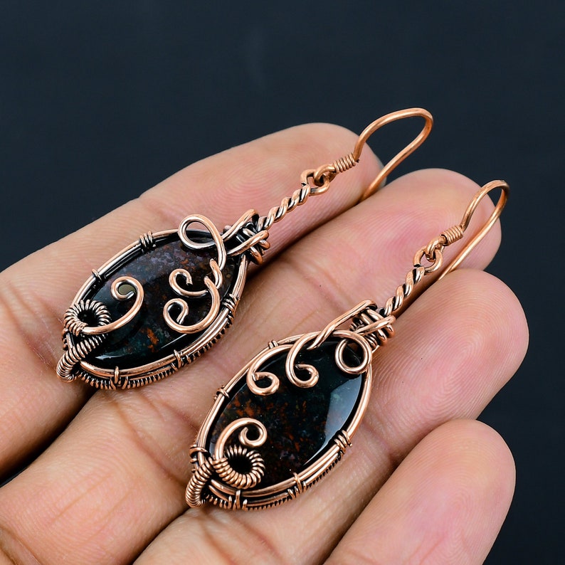May include: A pair of copper wire wrapped earrings with a black and brown oval shaped stone. The earrings have a delicate, intricate design.
