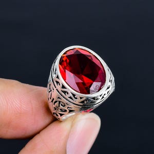 Peut inclure: Une bague en argent ornée d'une grande pierre précieuse rouge foncé de taille ovale sertie dans une monture. L'anneau présente des motifs en filigrane ajourés complexes qui s'enroulent sur les côtés, offrant un aspect texturé détaillé.