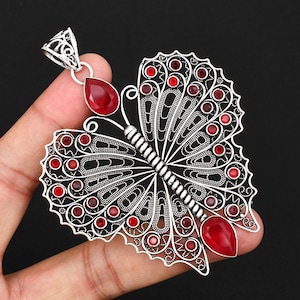 May include: A silver-toned butterfly pendant featuring intricate filigree metalwork and multiple small, round, red gemstones embedded throughout the wings. The pendant includes two larger, teardrop-shaped red stones at the top and bottom.