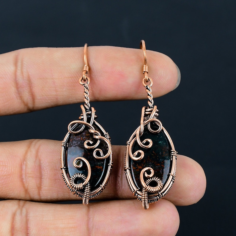 May include: A pair of copper wire wrapped earrings with oval shaped green and brown jasper stones. The earrings have a delicate, intricate design.