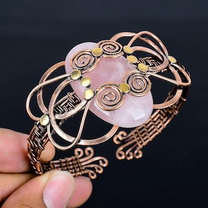May include: A copper-toned wire-wrapped bracelet featuring a large, oval rose quartz stone. The bracelet has intricate wirework with spiral and woven details, accented by small gold-colored embellishments. The open cuff design allows for easy wear.