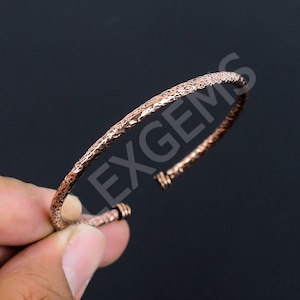 Peut inclure: Un bracelet manchette de couleur cuivre présentant une surface texturée et martelée avec un design fin et ouvert. La bande métallique possède une finition sombre et rustique avec des indentations visibles et des détails en fil enroulé aux extrémités.
