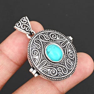 May include: A round silver-toned metal pendant featuring an intricate embossed scrollwork pattern and a central oval-shaped turquoise-coloured stone. The pendant includes a triangular-shaped bail at the top with matching decorative metal detailing.