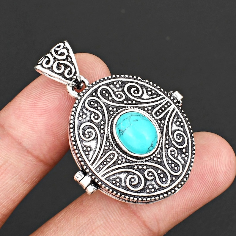 May include: A round silver-toned metal pendant featuring an intricate embossed scrollwork pattern and a central oval-shaped turquoise-coloured stone. The pendant includes a triangular-shaped bail at the top with matching decorative metal detailing.