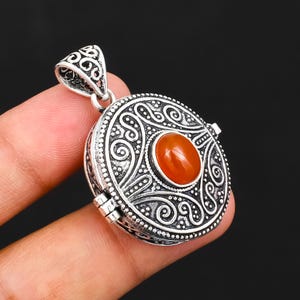 May include: A round silver locket pendant featuring intricate embossed scrollwork and a central oval-shaped orange carnelian gemstone. The pendant includes a decorative bail with matching filigree patterns and a visible side hinge mechanism.