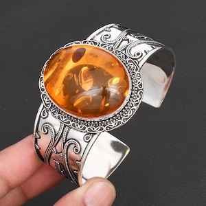 May include: Silver cuff bracelet featuring a large, oval amber stone. The amber is a warm, translucent orange color with natural inclusions. The silver band has an ornate, engraved design.