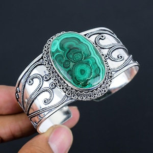 May include: A silver cuff bracelet featuring a large, oval-shaped green malachite gemstone with concentric banding patterns. The metal band is decorated with intricate scrollwork and beaded detailing surrounding the central stone setting.