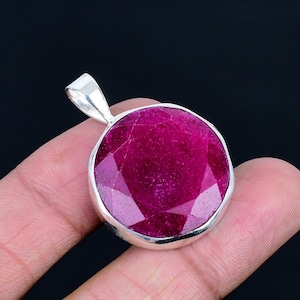 May include: A round, faceted, deep magenta-colored gemstone pendant set in a polished silver-toned metal bezel. The gemstone features a flat, multi-faceted surface that reflects light, and the pendant includes a silver-toned metal bail at the top.