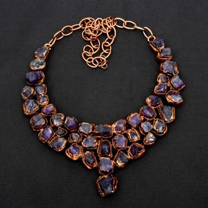 May include: A statement necklace featuring multiple raw, deep purple amethyst stones set in a copper-toned metal frame. The design consists of several layered clusters of jagged, unpolished gemstones connected by a copper-coloured chain link necklace.